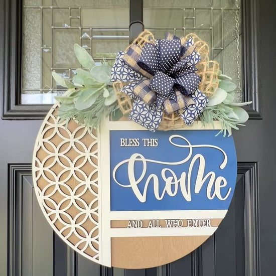 Navy blue and natural wood Bless This Home door hanger with layered 3D lettering, geometric overlay design, greenery accents, and designer bow displayed on a black front door.