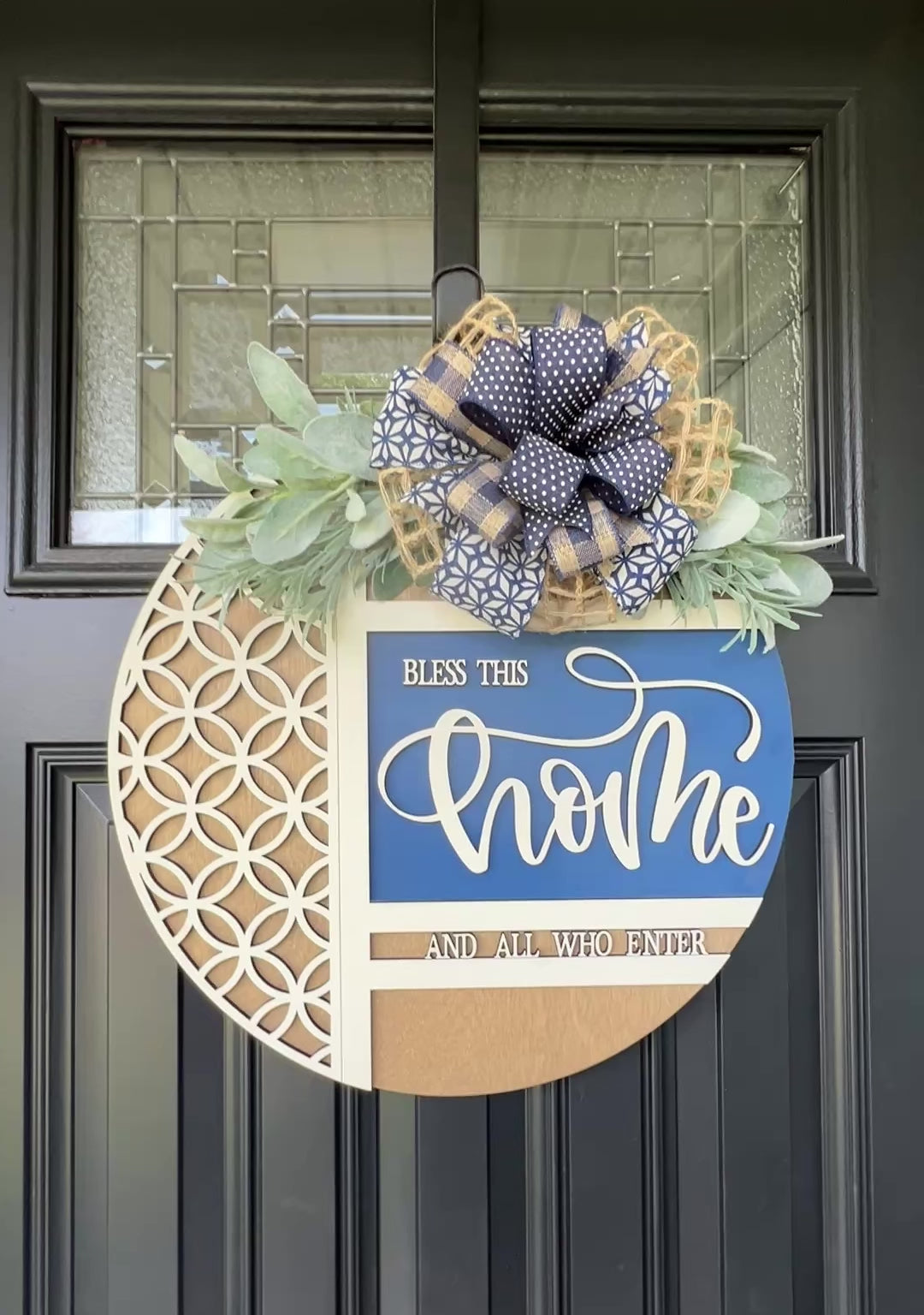Navy blue and natural wood Bless This Home door hanger with layered 3D lettering, geometric overlay design, greenery accents, and designer bow displayed on a black front door.
