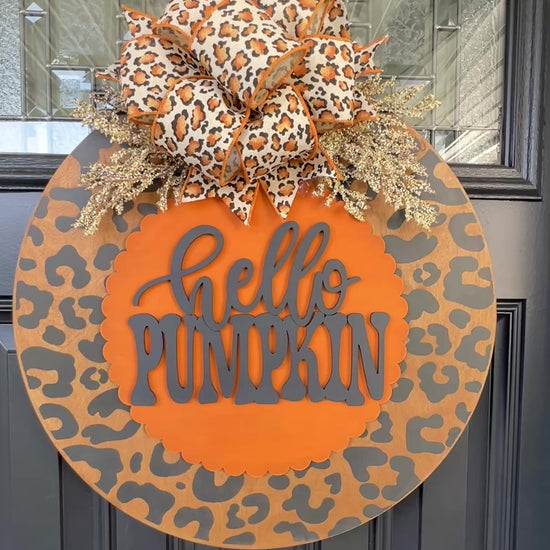 Hello Pumpkin leopard print fall door hanger with layered wood lettering, designer bow, seasonal greenery, and woodgrain animal print background displayed on a front door