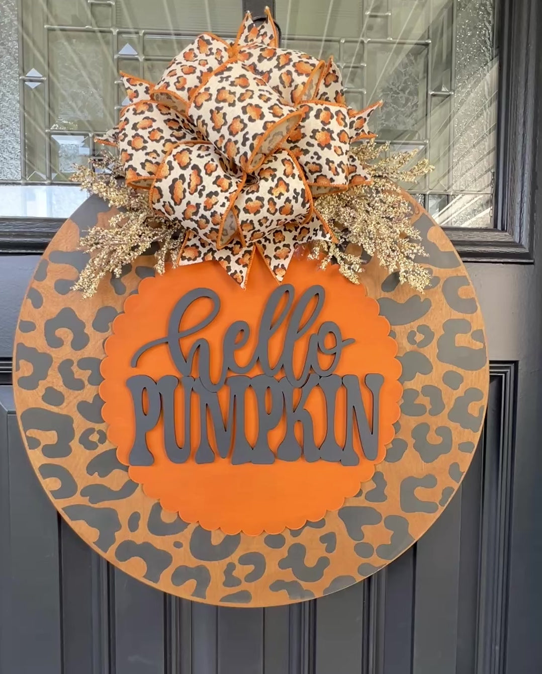 Hello Pumpkin leopard print fall door hanger with layered wood lettering, designer bow, seasonal greenery, and woodgrain animal print background displayed on a front door