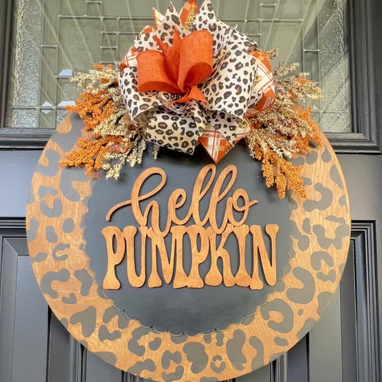 Hello Pumpkin leopard print fall door hanger with layered wood lettering, designer bow, seasonal greenery, and woodgrain animal print background displayed on a front door