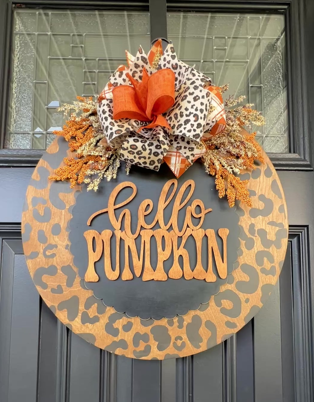 Hello Pumpkin leopard print fall door hanger with layered wood lettering, designer bow, seasonal greenery, and woodgrain animal print background displayed on a front door