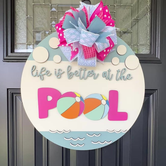 Handcrafted round door hanger featuring the phrase "Life Is Better at the Pool" with colorful beach ball accents, summer colors, and a bright pink and blue designer bow displayed on a front door.