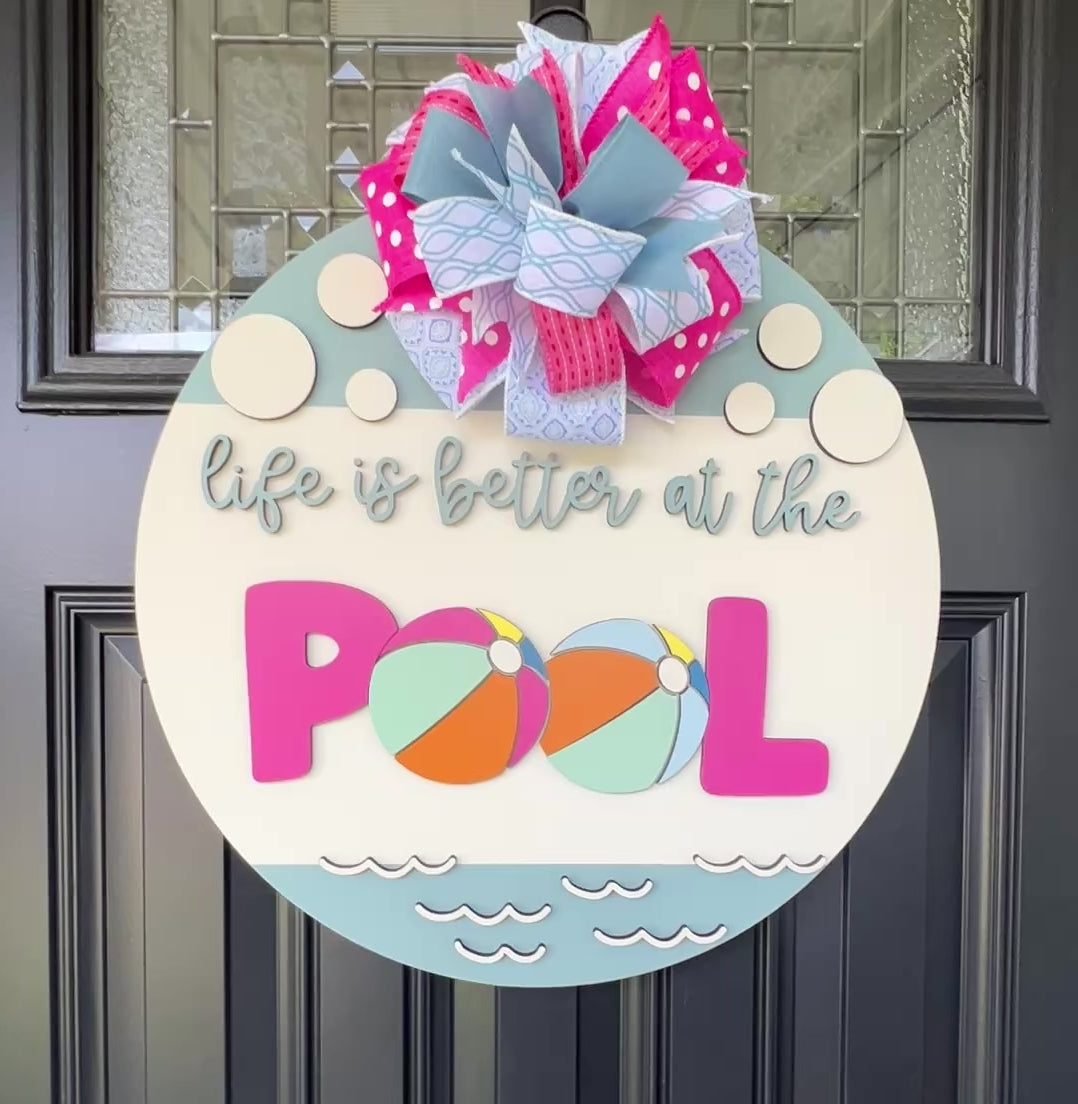 Handcrafted round door hanger featuring the phrase "Life Is Better at the Pool" with colorful beach ball accents, summer colors, and a bright pink and blue designer bow displayed on a front door.