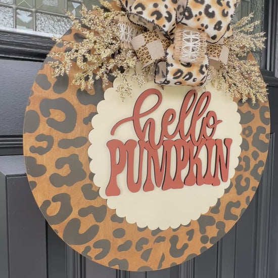 Hello Pumpkin leopard print fall door hanger with layered wood lettering, designer bow, seasonal greenery, and woodgrain animal print background displayed on a front door