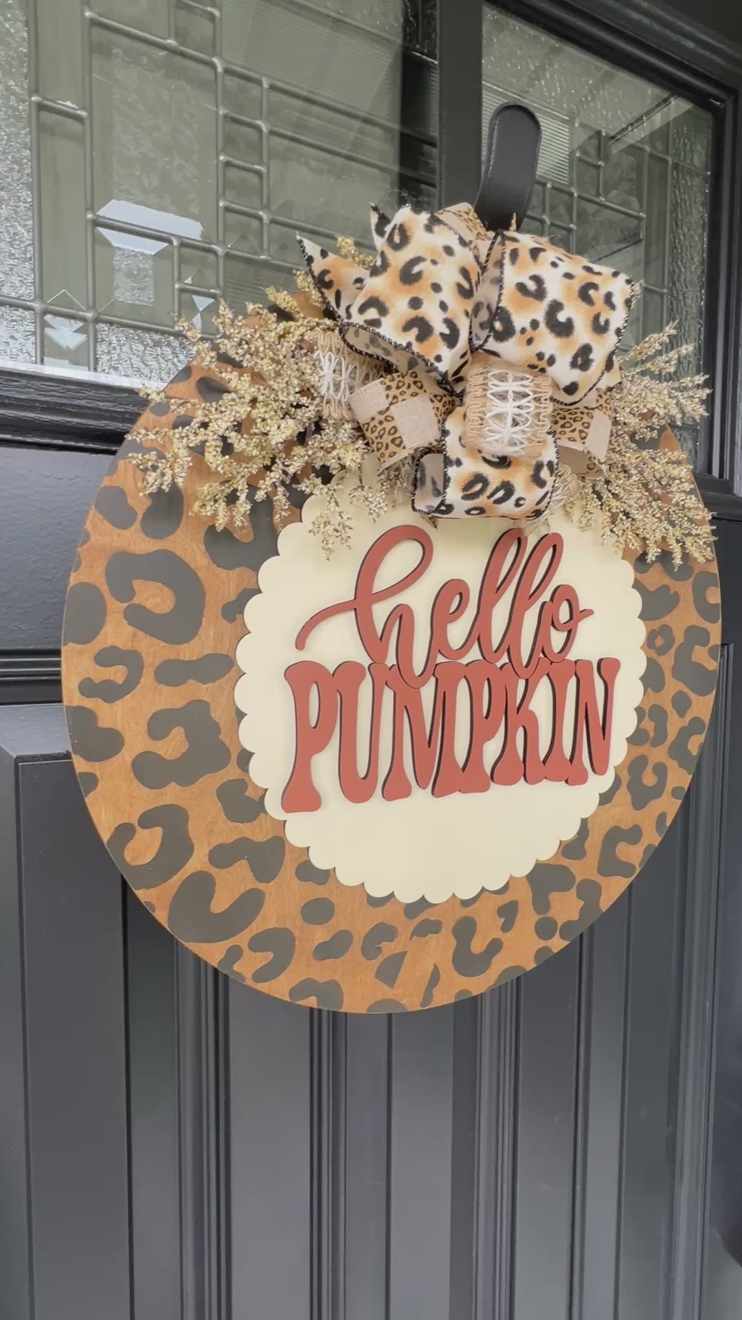 Hello Pumpkin leopard print fall door hanger with layered wood lettering, designer bow, seasonal greenery, and woodgrain animal print background displayed on a front door
