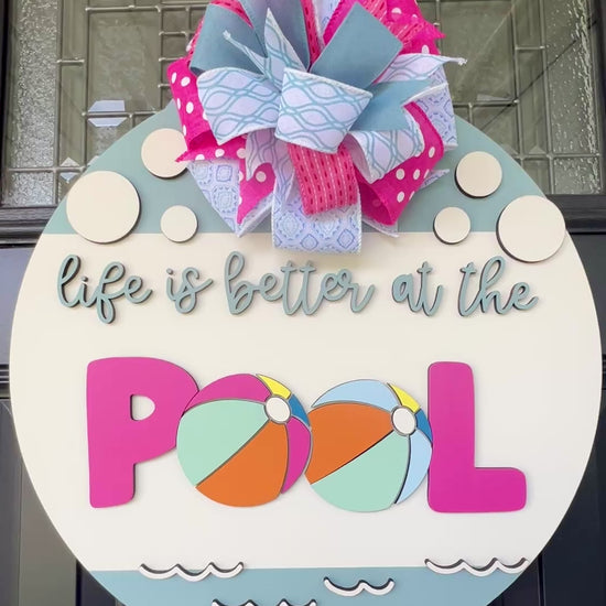 Handcrafted round door hanger featuring the phrase "Life Is Better at the Pool" with colorful beach ball accents, summer colors, and a bright pink and blue designer bow displayed on a front door.