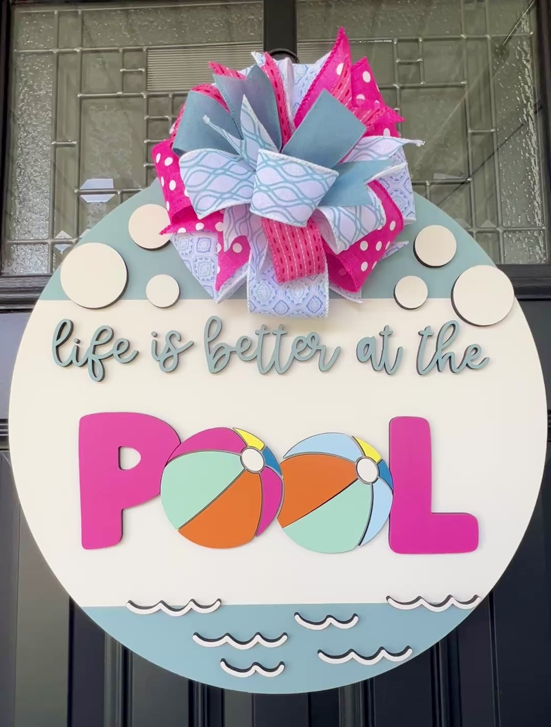 Handcrafted round door hanger featuring the phrase "Life Is Better at the Pool" with colorful beach ball accents, summer colors, and a bright pink and blue designer bow displayed on a front door.