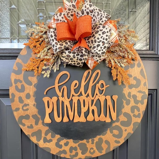 Hello Pumpkin leopard print fall door hanger with layered wood lettering, designer bow, seasonal greenery, and woodgrain animal print background displayed on a front door
