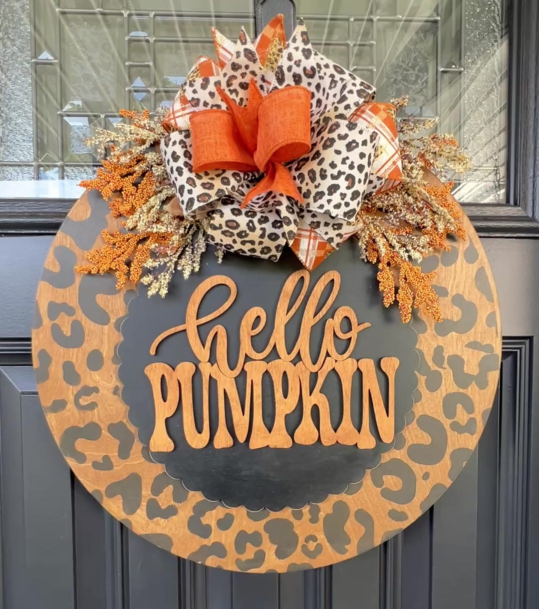Hello Pumpkin leopard print fall door hanger with layered wood lettering, designer bow, seasonal greenery, and woodgrain animal print background displayed on a front door