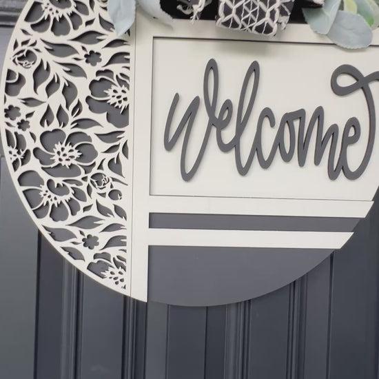 black and white welcome door hanger with floral cutout design
modern farmhouse front door sign with layered wood welcome script
18 inch round wooden welcome sign with decorative bow and greenery
neutral black white door hanger for front porch decor
handmade welcome sign with floral overlay and raised lettering
elegant entryway decor black and white round door hanger