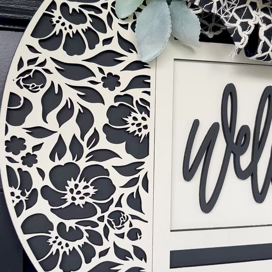 black and white welcome door hanger with floral cutout design
modern farmhouse front door sign with layered wood welcome script
18 inch round wooden welcome sign with decorative bow and greenery
neutral black white door hanger for front porch decor
handmade welcome sign with floral overlay and raised lettering
elegant entryway decor black and white round door hanger