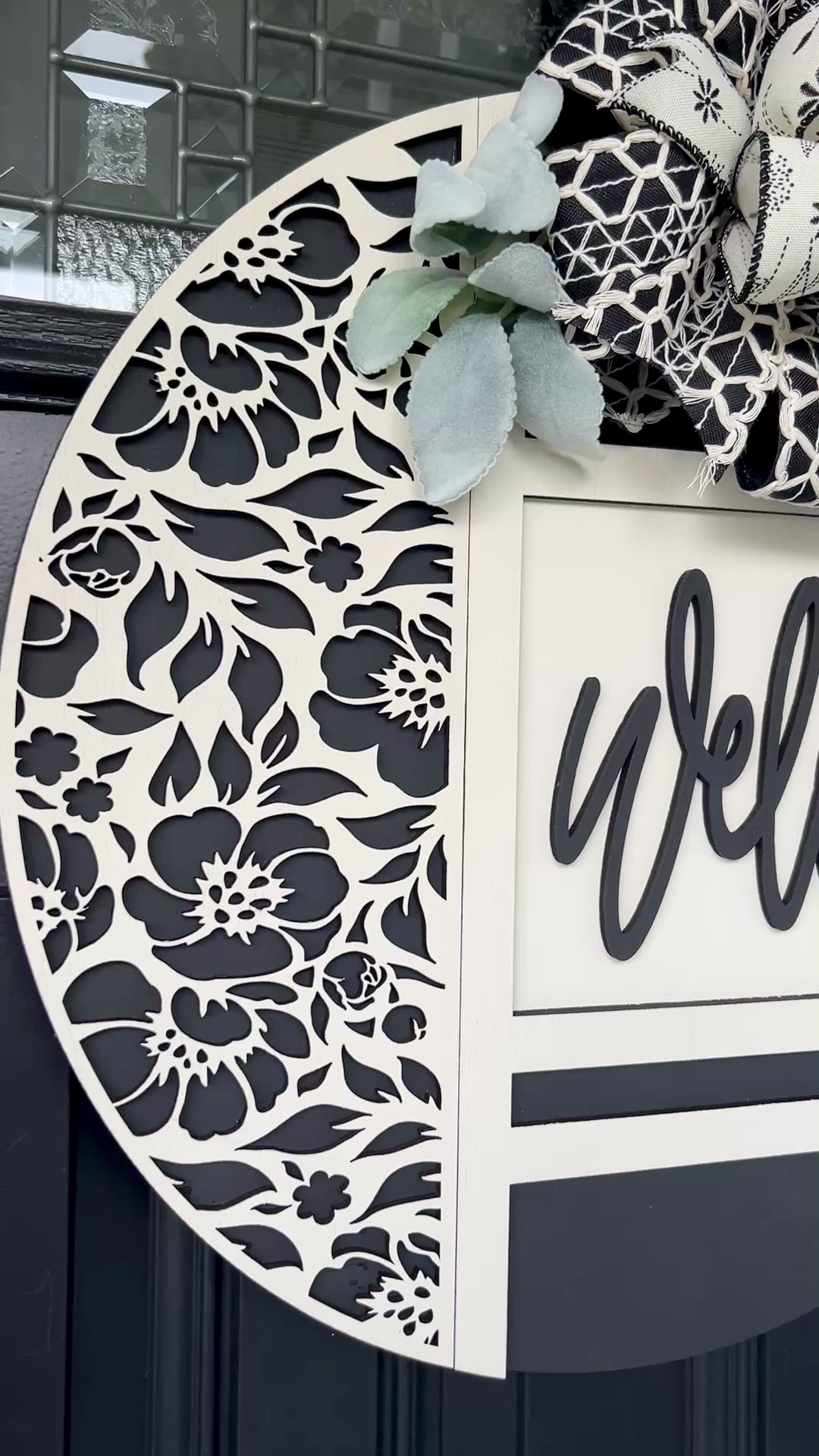 black and white welcome door hanger with floral cutout design
modern farmhouse front door sign with layered wood welcome script
18 inch round wooden welcome sign with decorative bow and greenery
neutral black white door hanger for front porch decor
handmade welcome sign with floral overlay and raised lettering
elegant entryway decor black and white round door hanger
