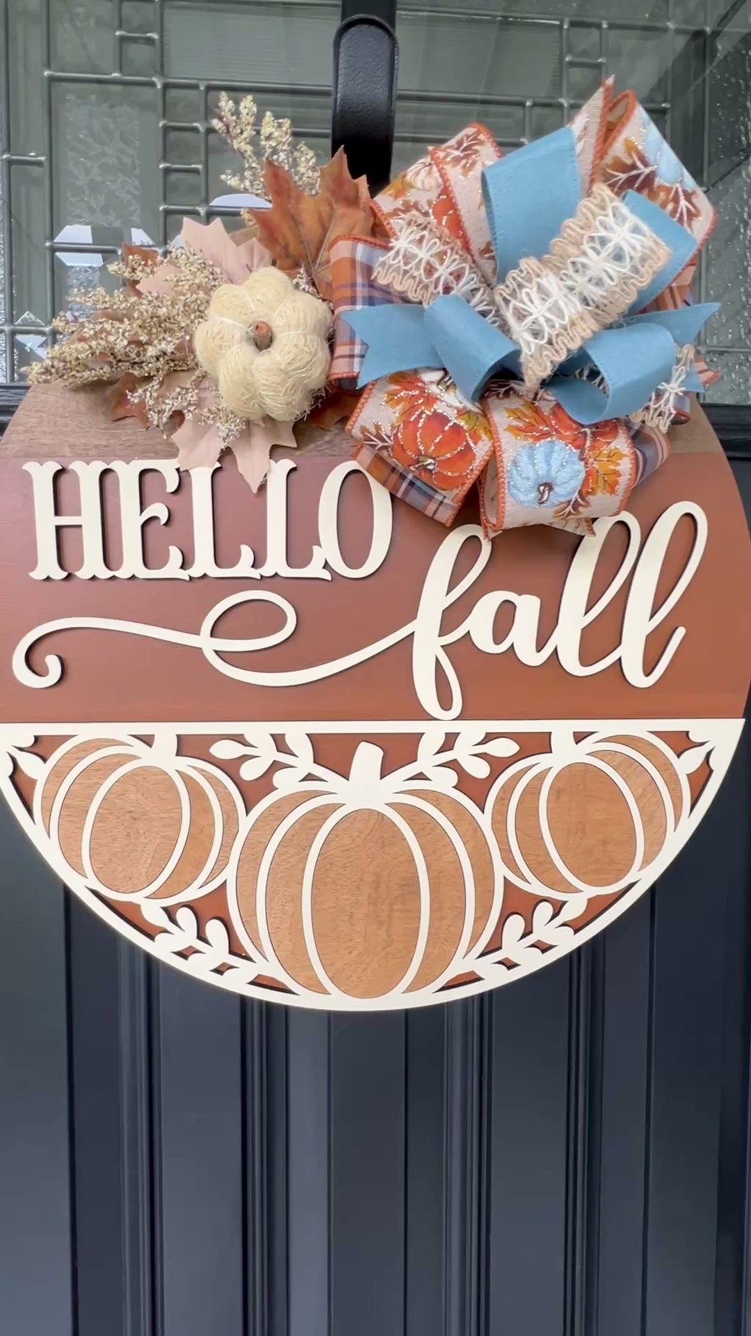 Hello Fall pumpkin door hanger with layered wood lettering, decorative pumpkin cutouts, designer bow, seasonal greenery, and autumn accents displayed on a front door.  Movie